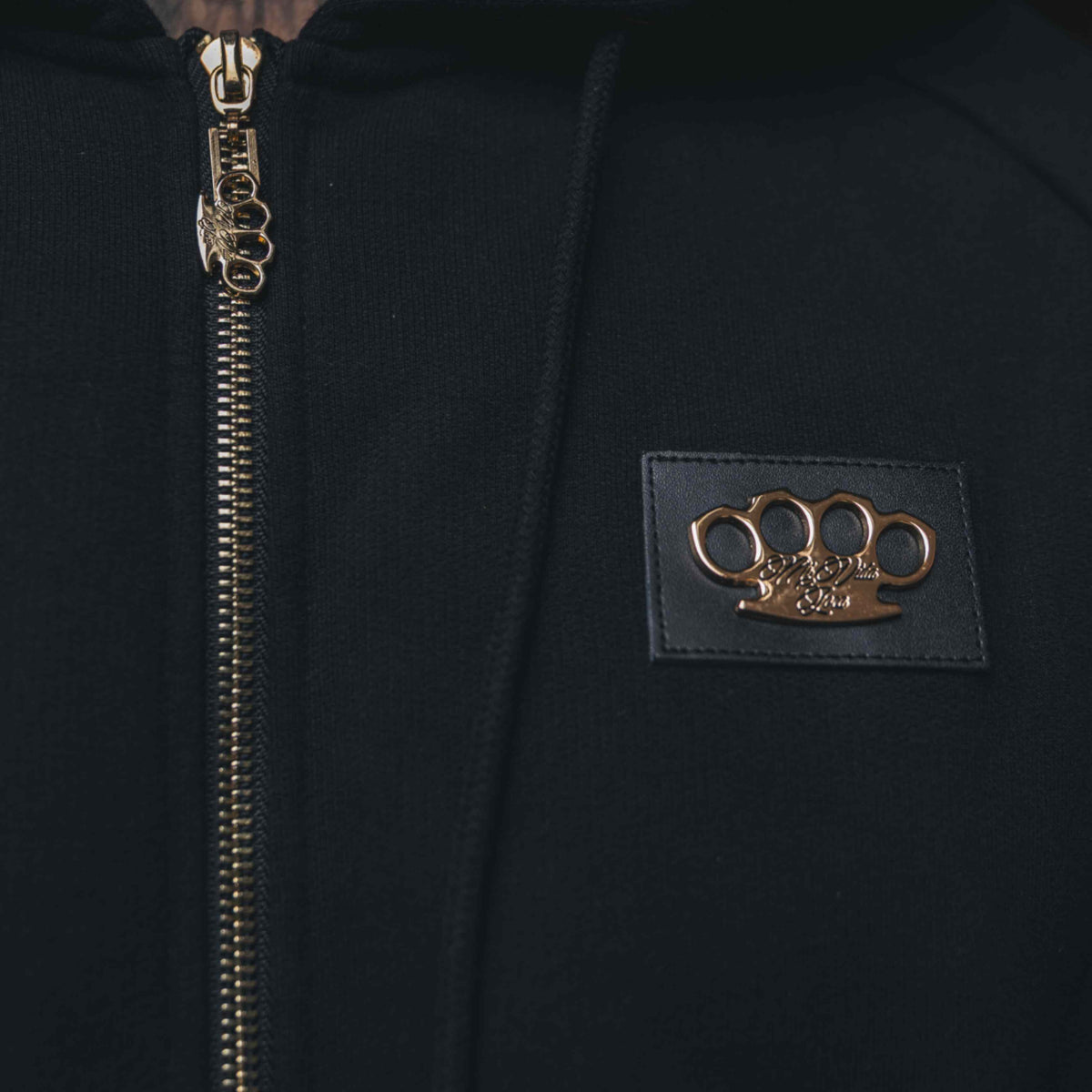 Gros plan du logo MVL doré sur patch en cuir et de la fermeture éclair dorée du hoodie Meander