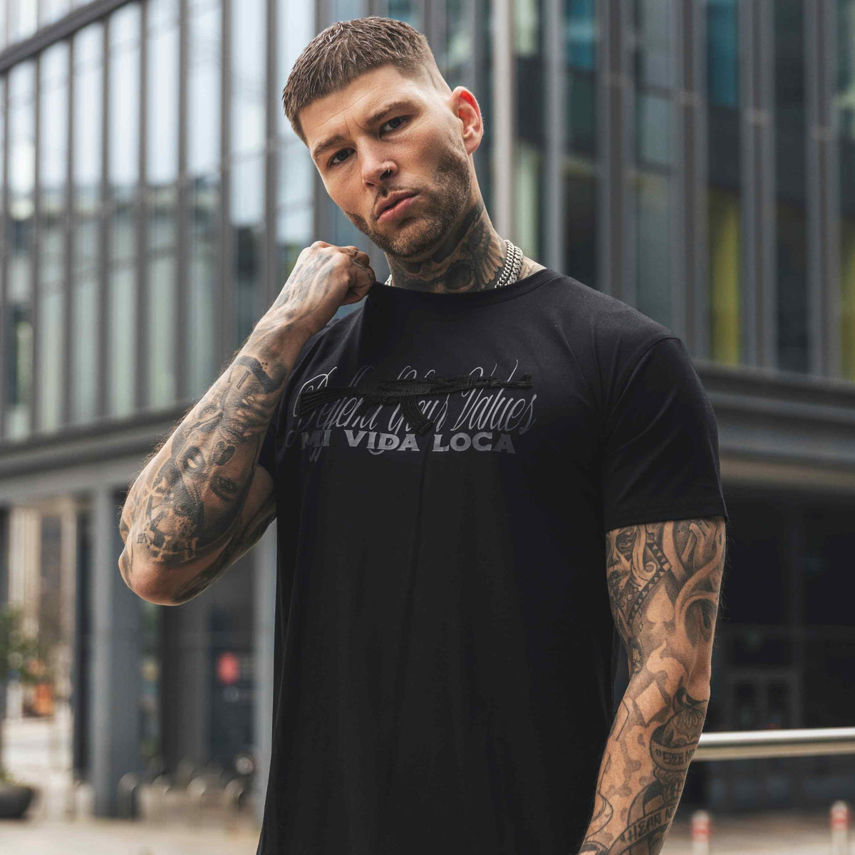Modelo con tatuajes lleva la camiseta MVL Defend your values negra delante de un edificio de cristal