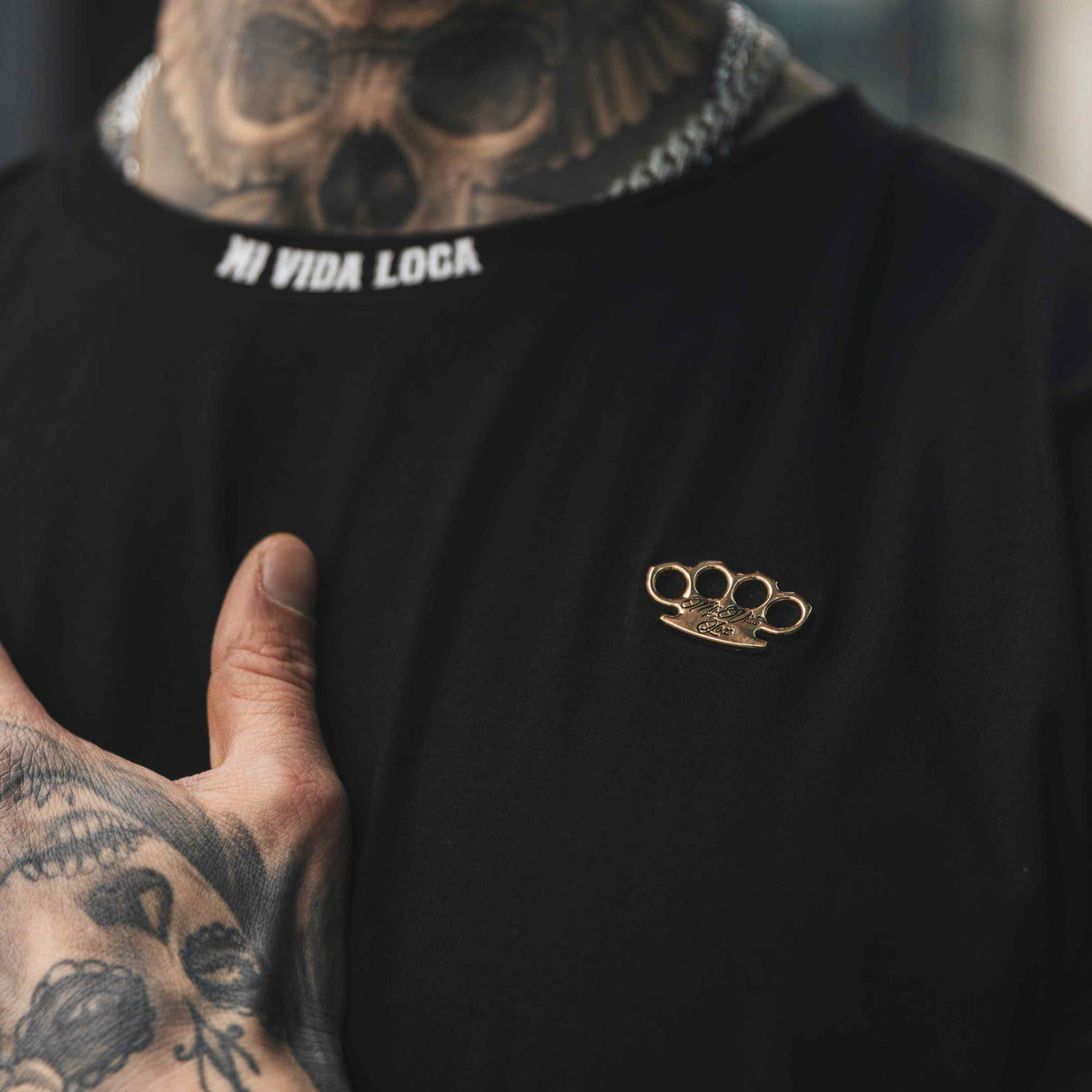 Nahaufnahme des goldenen MVL-Logos und des weißen Mi Vida Loca Schriftzugs im Kragen des schwarzen FTS T-Shirts