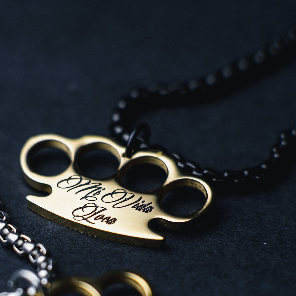 Close-up of the gold MVL logo pendant with engraved Mi Vida Loca lettering on the black chain